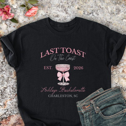 Black shirt with 'Last Toast on the Coast' design on a stone surface with flowers.