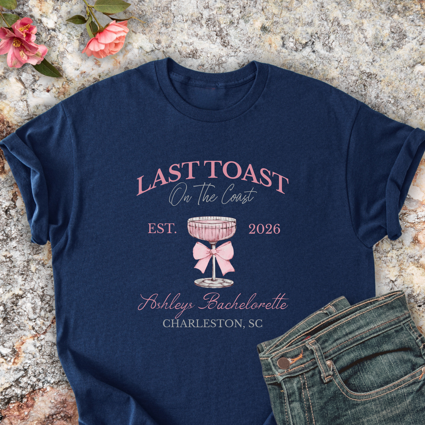 Navy shirt with 'Last Toast on the Coast' design on a stone surface with flowers.