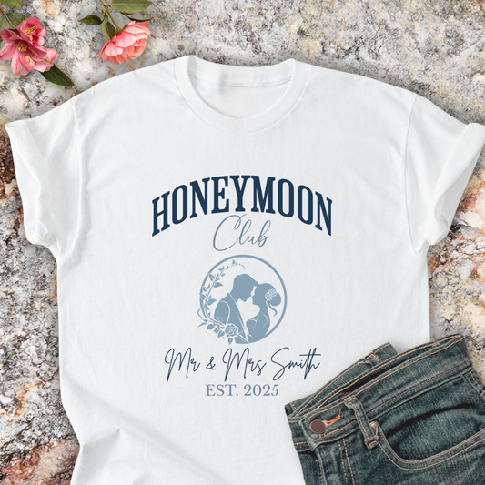 White shirt with 'Honeymoon Club' design on a textured surface with flowers and jeans.