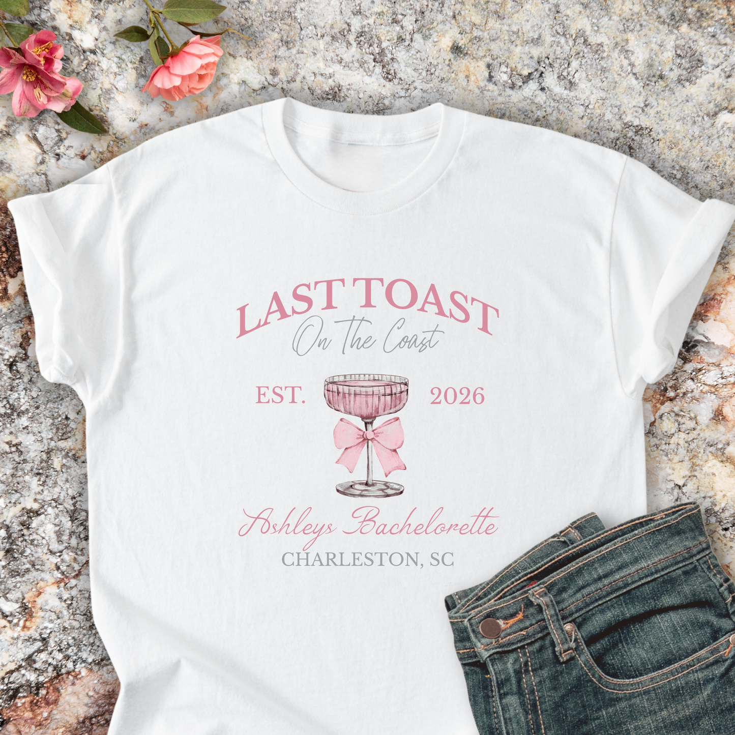 White shirt with 'Last Toast on the Coast' design on a stone surface with flowers.