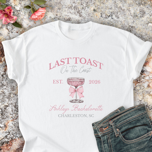 White shirt with 'Last Toast on the Coast' design on a stone surface with flowers.
