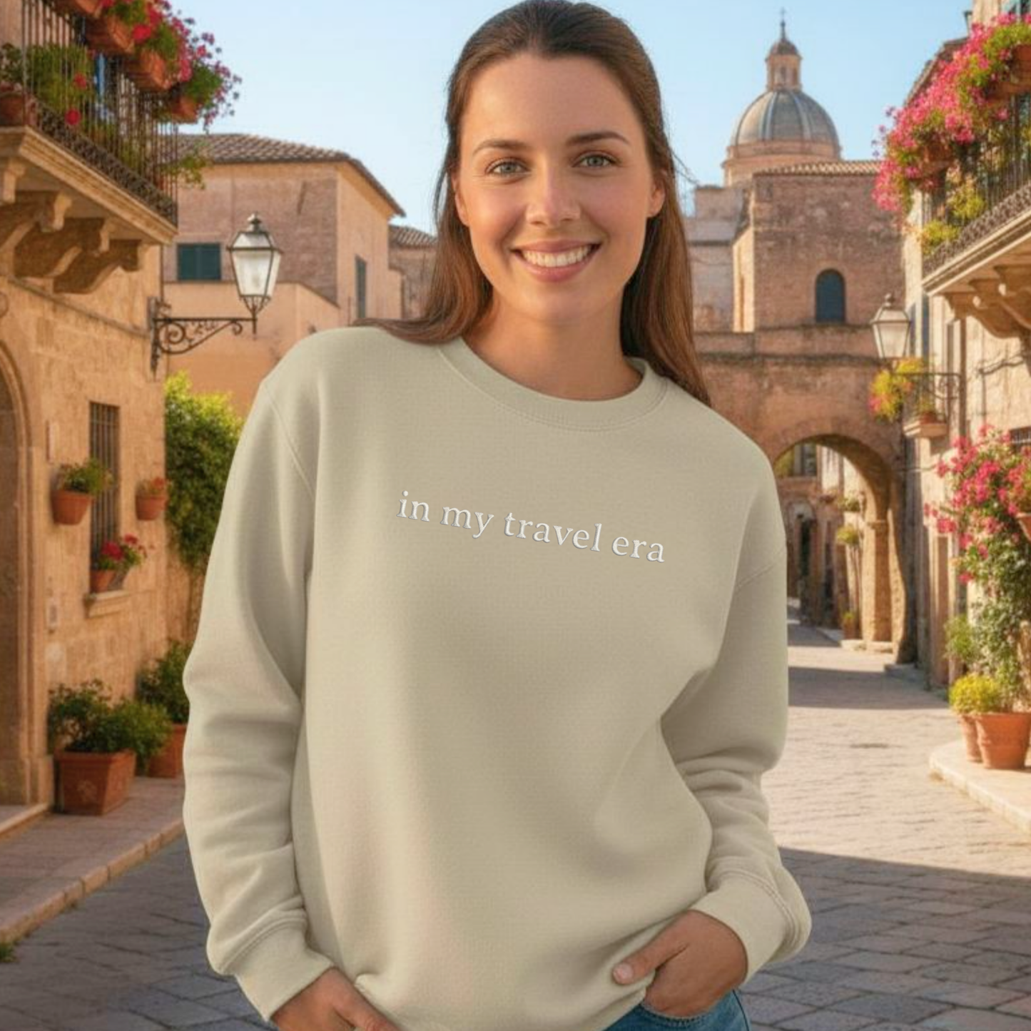 Woman wearing a sand sweatshirt with 'in my travel era' text, standing in a town street.
