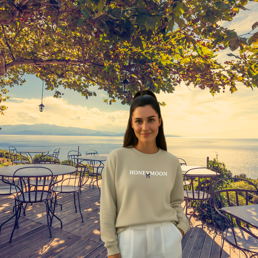 Woman wearing a 'Honeymoon' sweatshirt standing on a wooden deck with a scenic view of water and mountains.