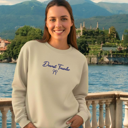 Woman wearing a sand sweatshirt with 'Dearest Traveler' text in front of a scenic lake and mountains.