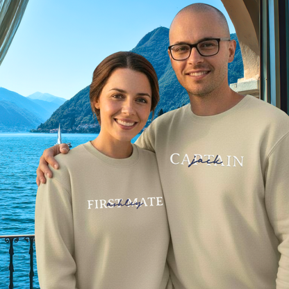 Two people wearing "FIRST MATE' and 'CAPTAIN' text sweatshirts, standing by a scenic lake with mountains.