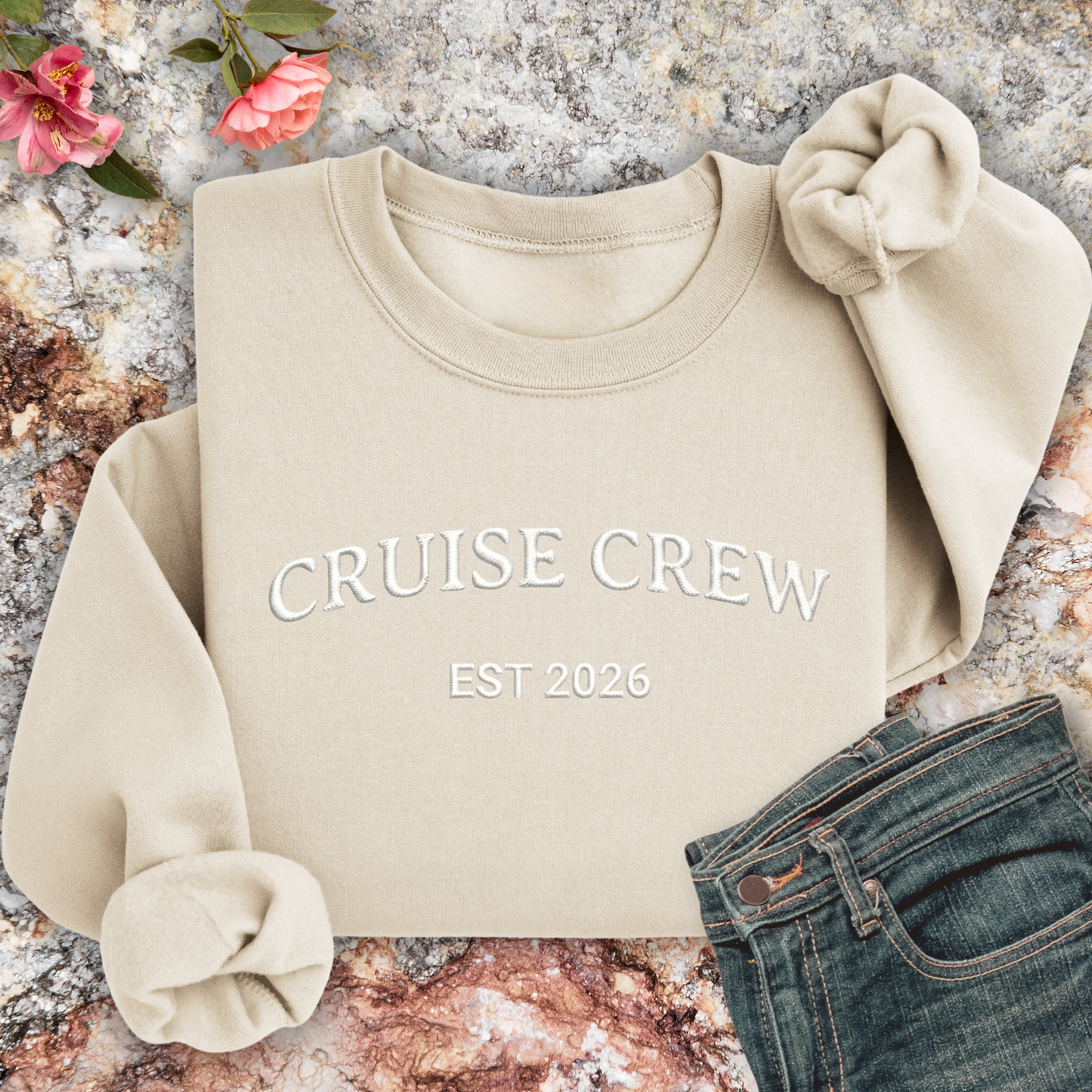Sand sweatshirt with 'CRUISE CREW EST 2026' text on a textured surface with flowers and jeans.