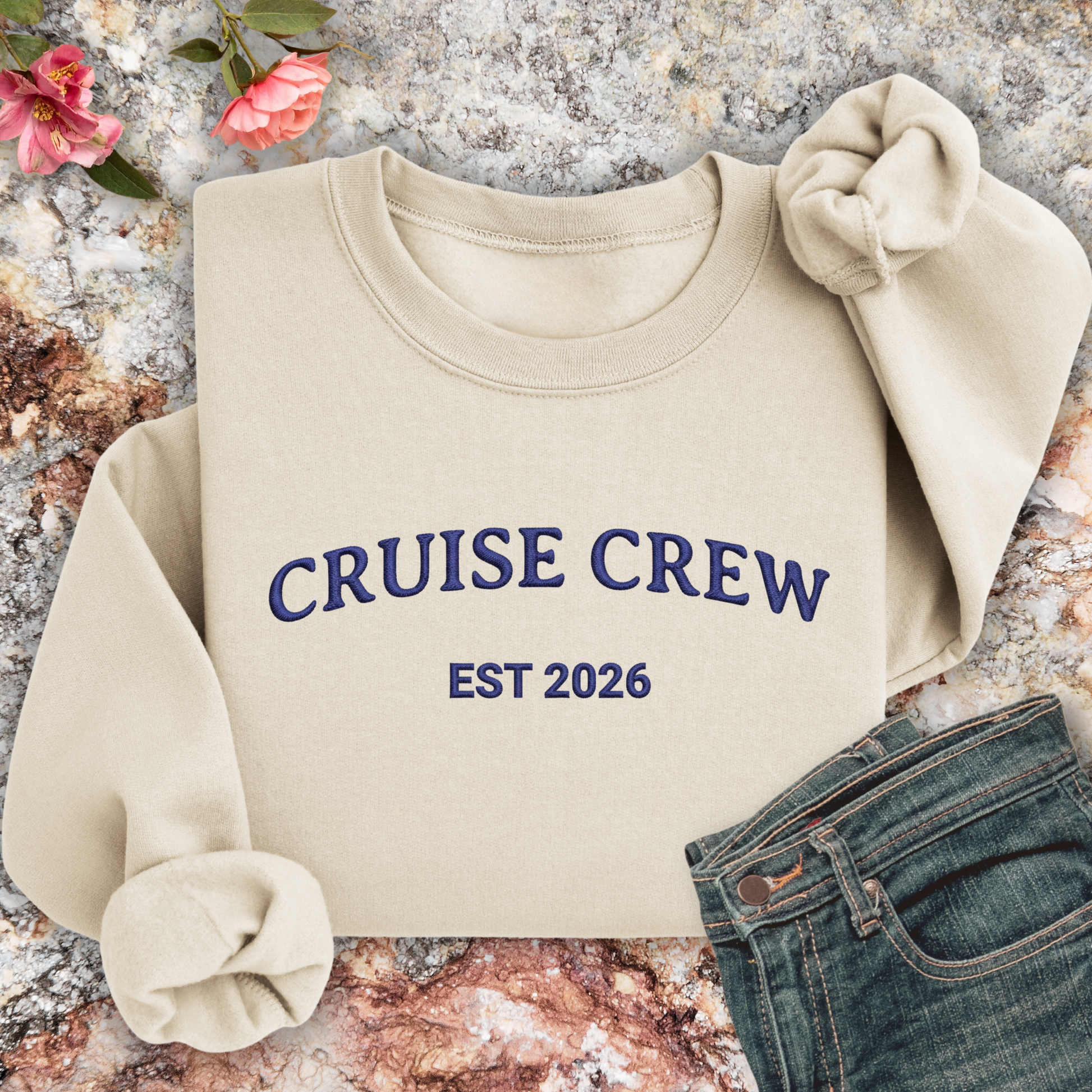 Sand sweatshirt with 'CRUISE CREW EST 2026' text on a textured surface with flowers and jeans.