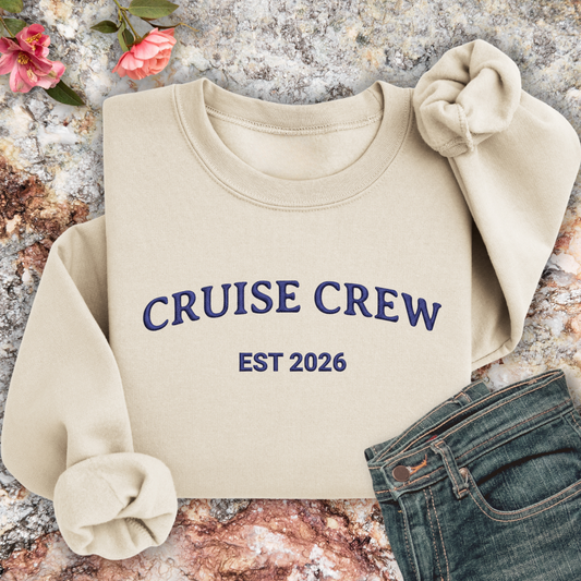 Sand sweatshirt with 'CRUISE CREW EST 2026' text on a textured surface with flowers and jeans.