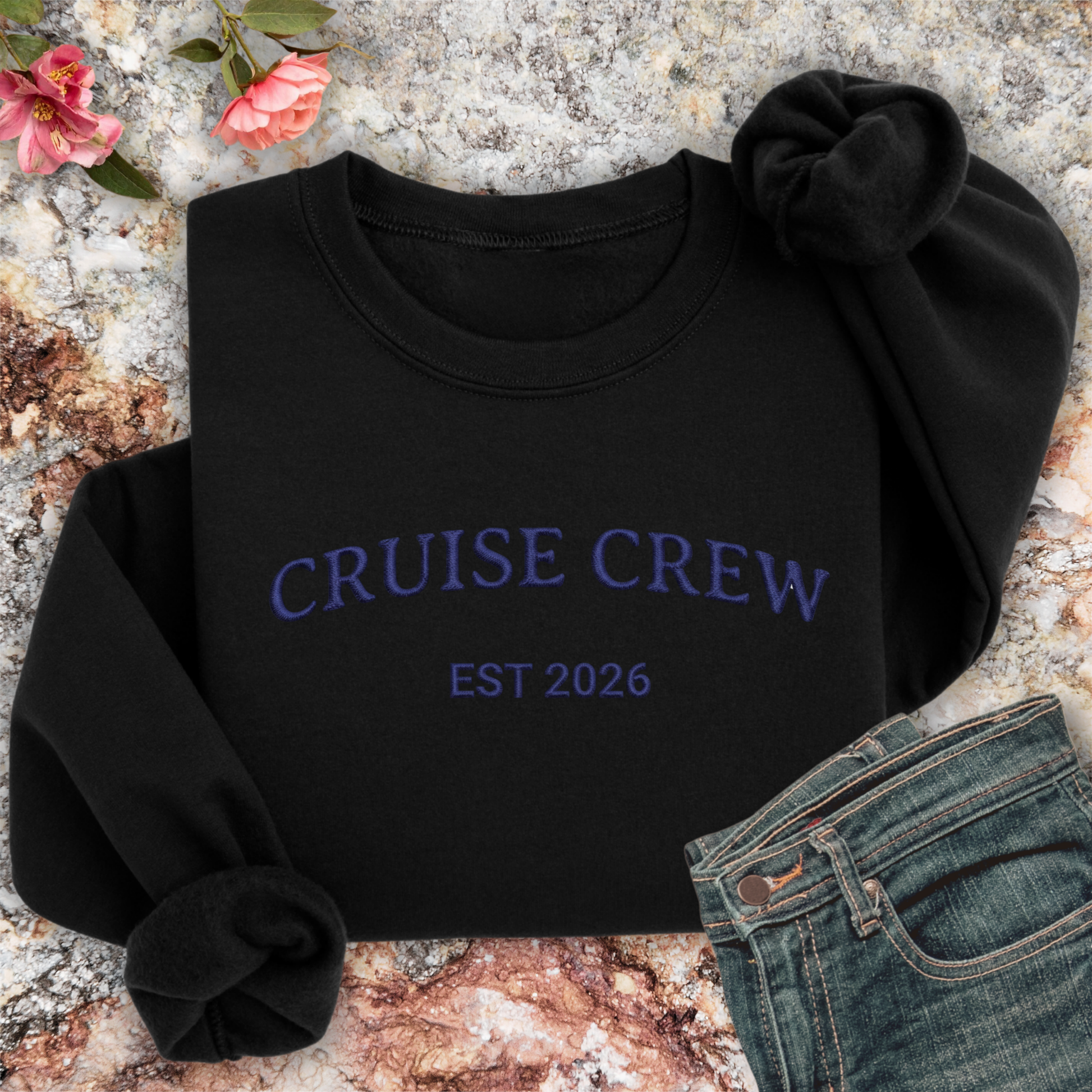 Black sweatshirt with 'CRUISE CREW EST 2026' text on a textured surface with flowers and jeans.