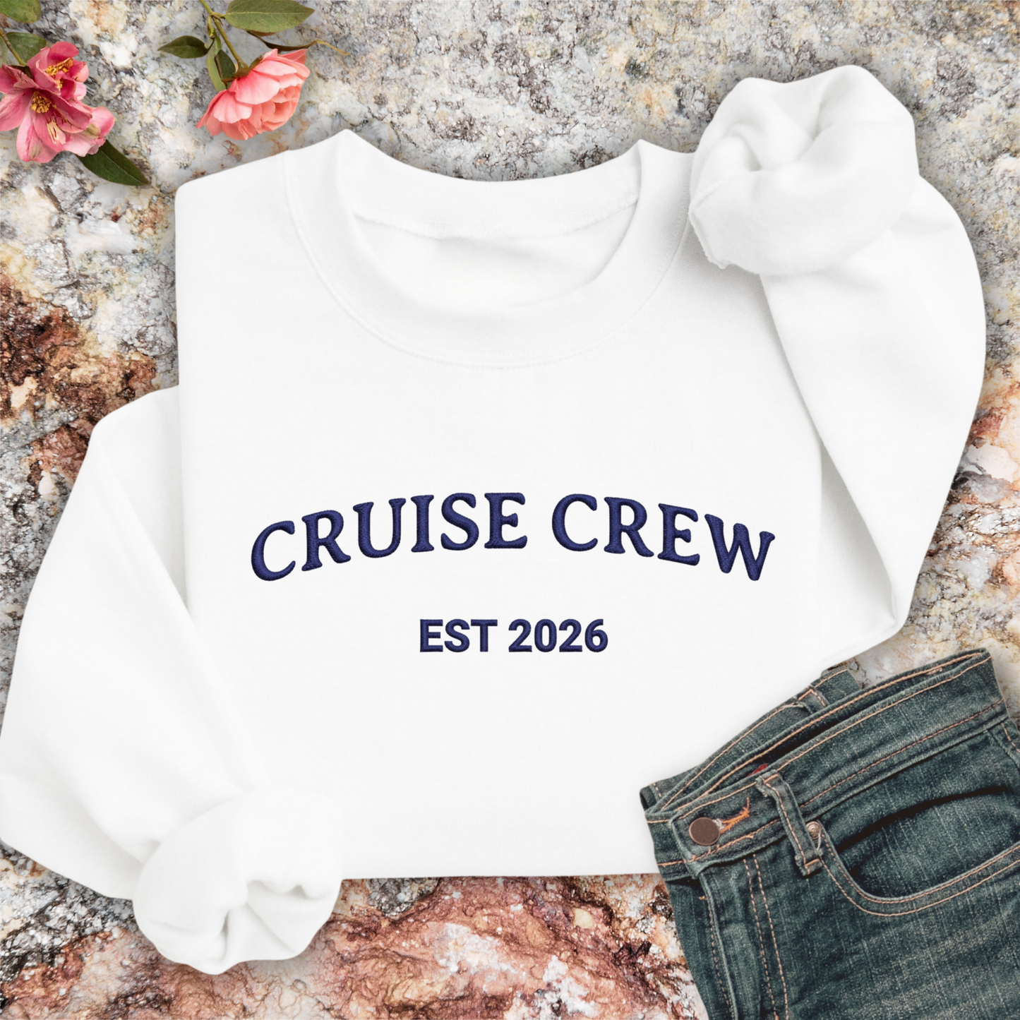 White sweatshirt with 'CRUISE CREW EST 2026' text on a textured surface with flowers and jeans.