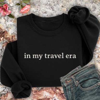 Black sweatshirt with 'in my travel era' text on a textured surface with flowers and jeans.