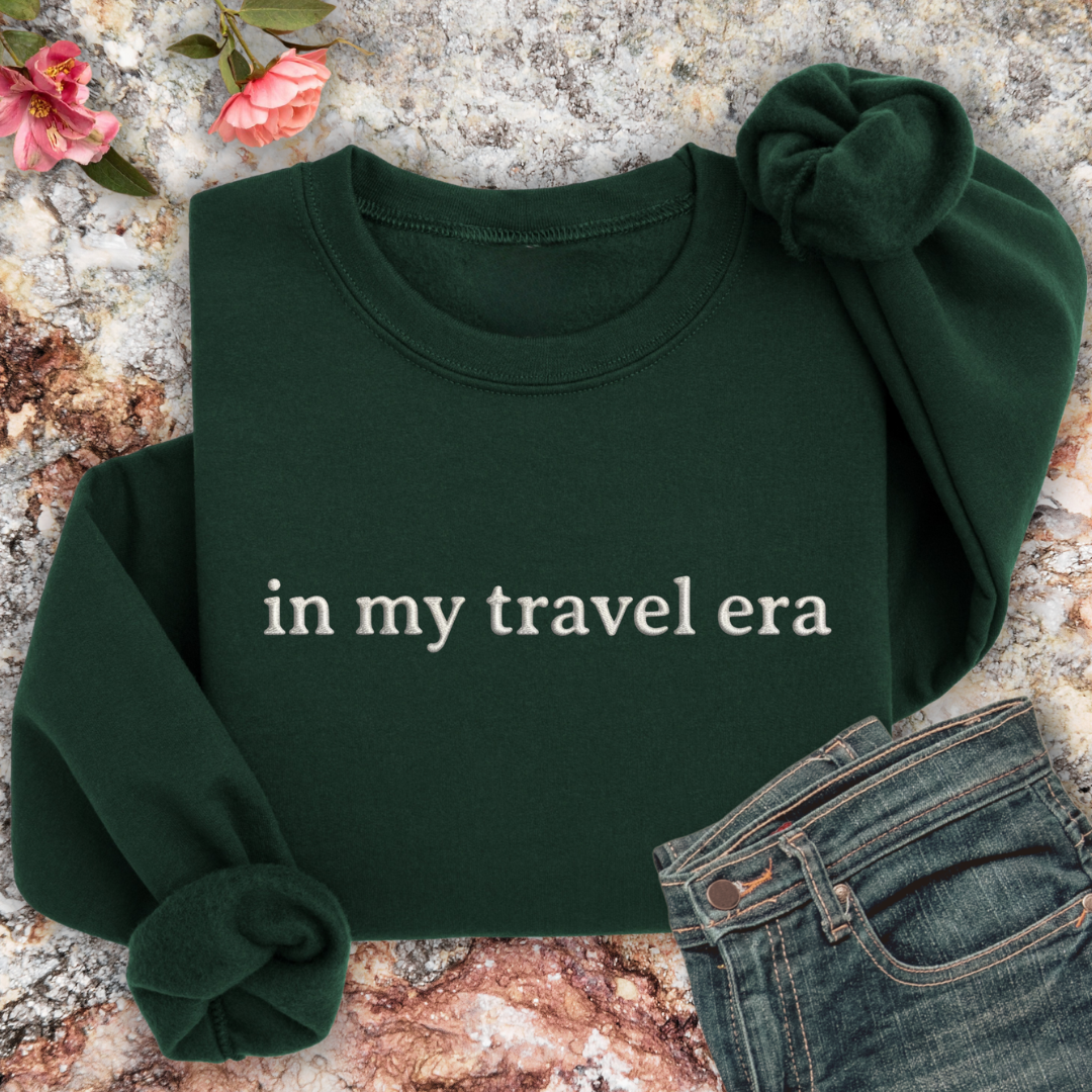 Forest Green sweatshirt with 'in my travel era' text on a textured surface with flowers and jeans.