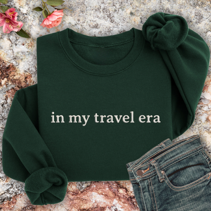 Forest Green sweatshirt with 'in my travel era' text on a textured surface with flowers and jeans.