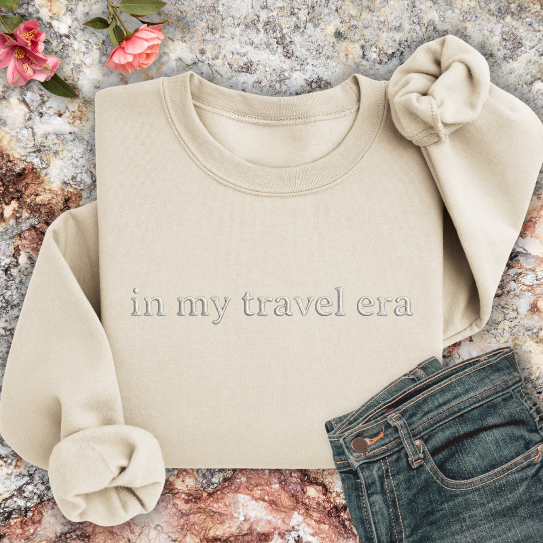 Sand sweatshirt with 'in my travel era' text on a textured surface with flowers and jeans.