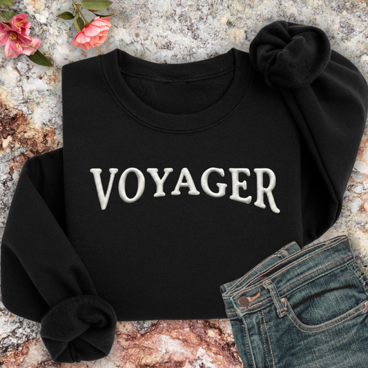Black sweatshirt with 'VOYAGER' text on a textured surface with flowers and jeans.